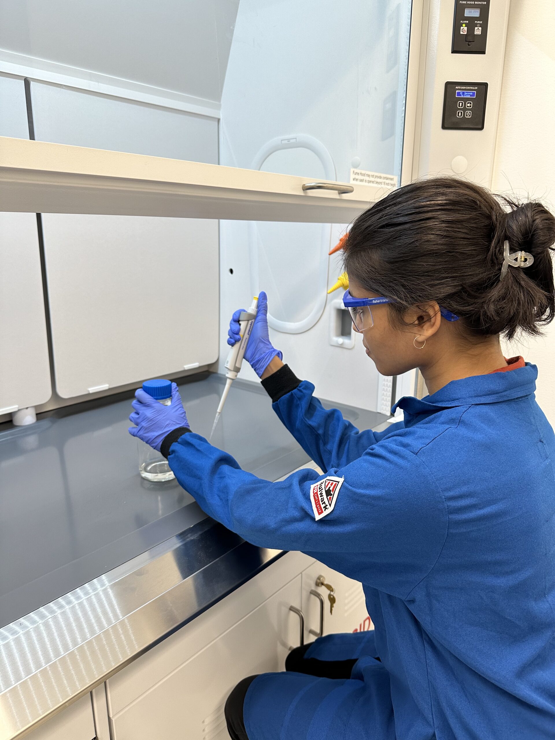 A women sits at a fume hood, pipetting liquid into a beaker.