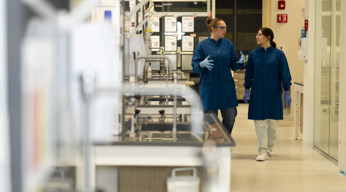 Two students in blue lab coats walk through a laboratory talking to one another.