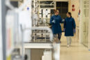 Two students in blue lab coats walk through a laboratory talking to one another.